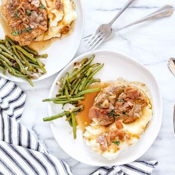 Saucy Pork Chops with Prosciutto. Overview of 2 white round plate with Mashed Potato topped with a pork chop, prosciutto and sauce and green beans on the side of the mashed potatoes. There are two forks and a black and with striped towel. www.atwistedplate/saucy-pork-chops/